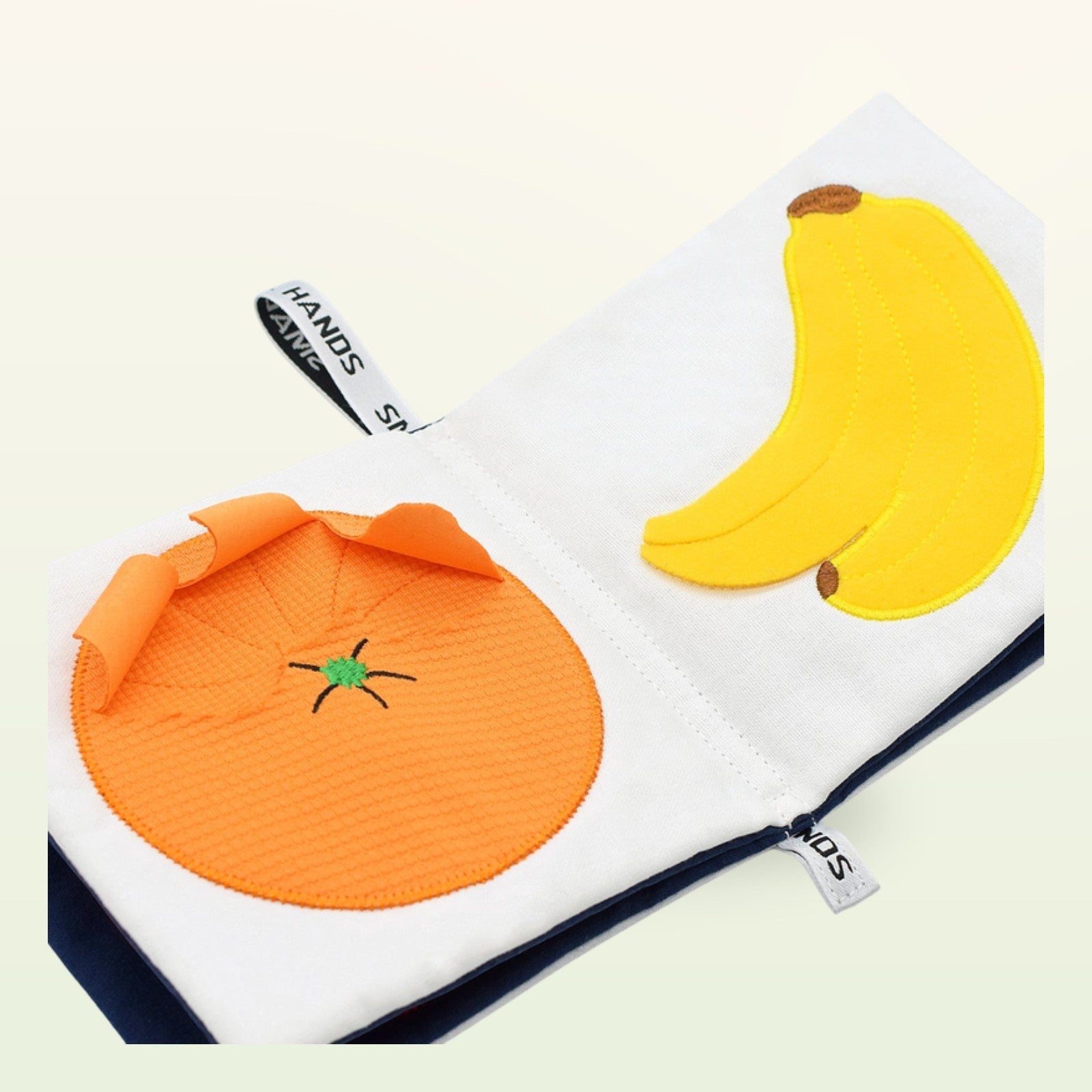 Small Hands Exotische Früchte Montessori Stoffbuch aufgeschlagen – Orange und Banane in Schrägansicht auf sanftem Creme-Hintergrund – ab 0 Monaten