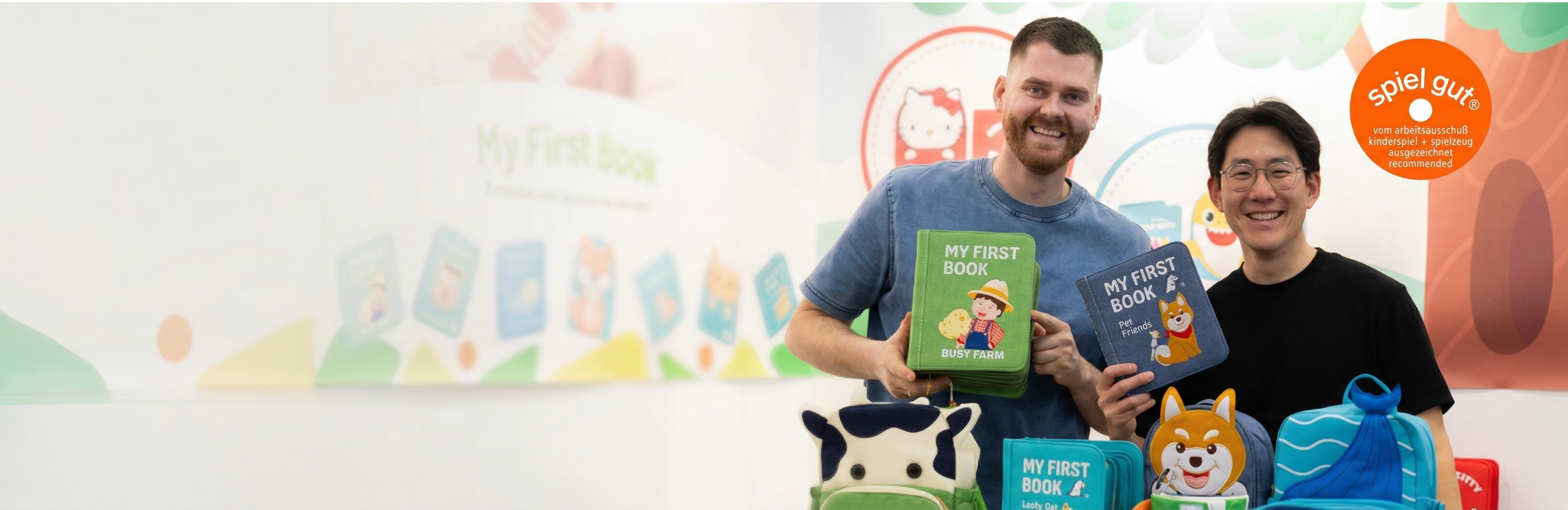 Jona the founder showcasing the bestseller My First Book Busy Farm and Pet Friends at the exhibition Babini. They are smiling and are happy. Also various products from My First Book as well as a Spiel Gut Zertifikat can be seen.